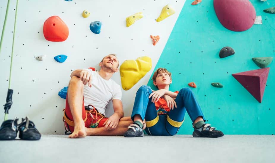 Father and teenage son sitting near the indoor climbing wall. They resting after the active climbing. Happy parenting concept image.